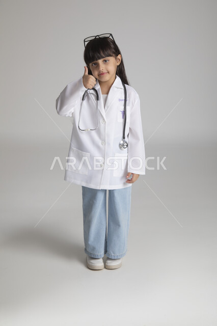 استمتاع الأطفال بالتجارب العملية للمهن المستقبلية، السعي نحو الأحلام والطموحات للدخول في مجال الطب والرعاية الصحية، بورتريه لبنت عربية خليجية سعودية ترتدي المعطف الطبي وتضع سماعة الفحص ترفع اصبعها بسرور بعلامة الجودة والقبول، الجسم بالطول الكامل، خلفية رمادية