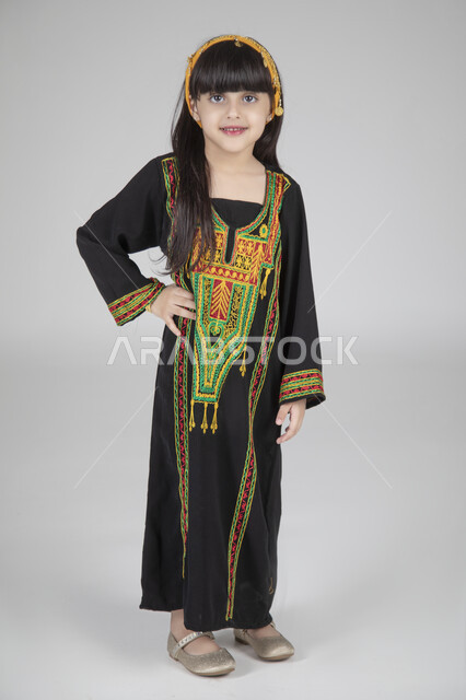 الاحتفال بيوم التأسيس والمناسبات الوطنية الشعبية، بورتريه لبنت عربية خليجية سعودية ترتدي الزي المطرز التقليدي السعودي النسائي تقف وتنظر إلى الكاميرا بإيماءات العز والفخر، ارتداء الأزياء التراثية القديمة، الجسم بالطول الكامل، خلفية رمادية