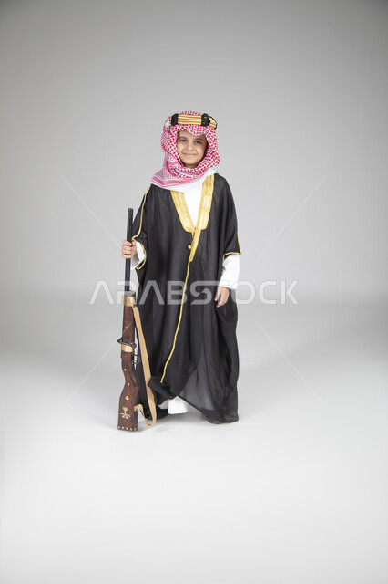 الاحتفال بالمناسبات الوطنية الشعبية، ارتداء الأزياء التقليدية التراثية في يوم التأسيس، بورتريه لولد عربي خليجي سعودي يرتدي البشت الأسود والعقال الذهبي العريض يقف ويحمل البندقية السعودية و ينظر إلى الكاميرا بإيماءات البشاشة، الجسم بالطول الكامل، خلفية رمادية