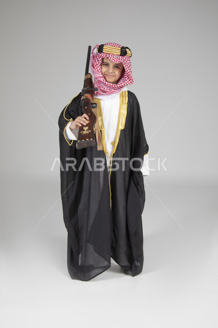 ارتداء الأزياء التقليدية التراثية في يوم التأسيس، الاحتفال بالمناسبات الوطنية الشعبية، بورتريه لولد عربي خليجي سعودي يرتدي البشت الأسود والعقال الذهبي العريض يقف ويحمل البندقية السعودية و ينظر إلى الكاميرا بإيماءات البشاشة، الجسم بالطول الكامل، خلفية رمادية