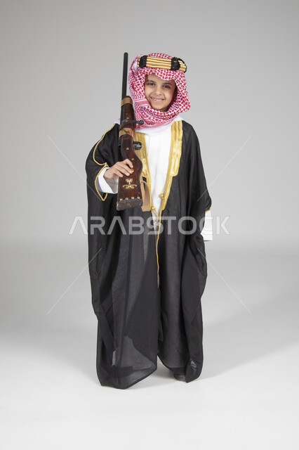 ارتداء الأزياء التقليدية التراثية في يوم التأسيس، الاحتفال بالمناسبات الوطنية الشعبية، بورتريه لولد عربي خليجي سعودي يرتدي البشت الأسود والعقال الذهبي العريض يقف ويحمل البندقية السعودية و ينظر إلى الكاميرا بإيماءات البشاشة، الجسم بالطول الكامل، خلفية رمادية