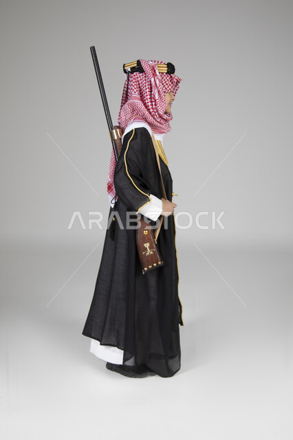 ارتداء الأزياء التقليدية التراثية في يوم التأسيس، الاحتفال بالمناسبات الوطنية الشعبية، بورتريه من الجانب لولد عربي خليجي سعودي يرتدي البشت الأسود والعقال الذهبي العريض يحمل البندقية السعودية وينظر إلى مكان ما بعيد، الجسم بالطول الكامل، خلفية رمادية