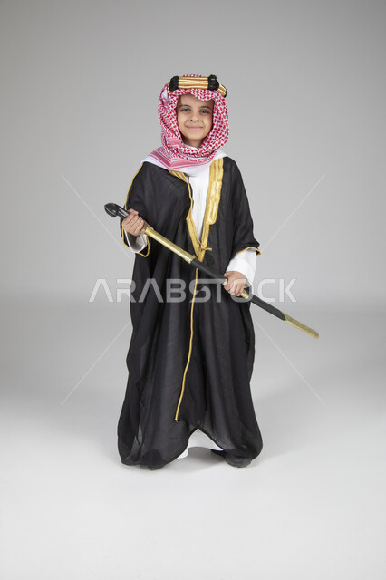  إحياء ذكرى يوم التأسيس والاحتفال بالمناسبات الوطنية الشعبية، بورتريه لولد عربي خليجي سعودي يرتدي البشت الأسود والعقال الذهبي العريض يمسك السيف بيده وينظر إلى الكاميرا بإيماءات العز والفخر، ارتداء الأزياء التقليدية التراثية، الجسم بالطول الكامل، خلفية رمادية