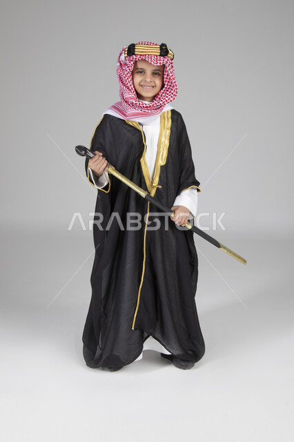  إحياء ذكرى يوم التأسيس والاحتفال بالمناسبات الوطنية الشعبية، بورتريه لولد عربي خليجي سعودي يرتدي البشت الأسود والعقال الذهبي العريض يمسك السيف بيده وينظر إلى الكاميرا بإيماءات العز والفخر، ارتداء الأزياء التقليدية التراثية، الجسم بالطول الكامل، خلفية رمادية