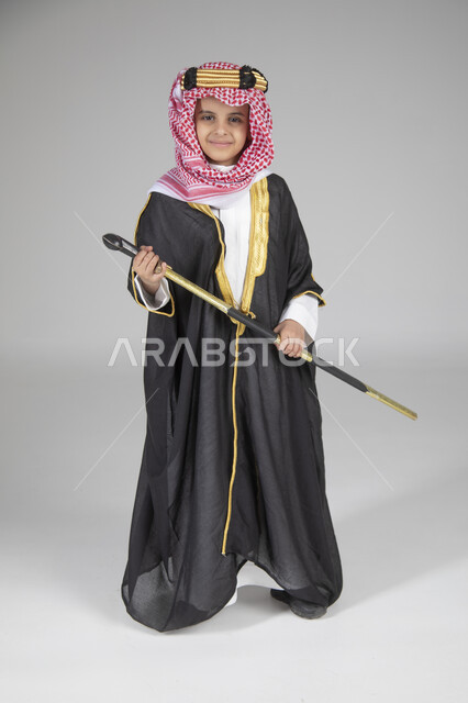  إحياء ذكرى يوم التأسيس والاحتفال بالمناسبات الوطنية الشعبية، بورتريه لولد عربي خليجي سعودي يرتدي البشت الأسود والعقال الذهبي العريض يمسك السيف بيده وينظر إلى الكاميرا بإيماءات العز والفخر، ارتداء الأزياء التقليدية التراثية، الجسم بالطول الكامل، خلفية رمادية