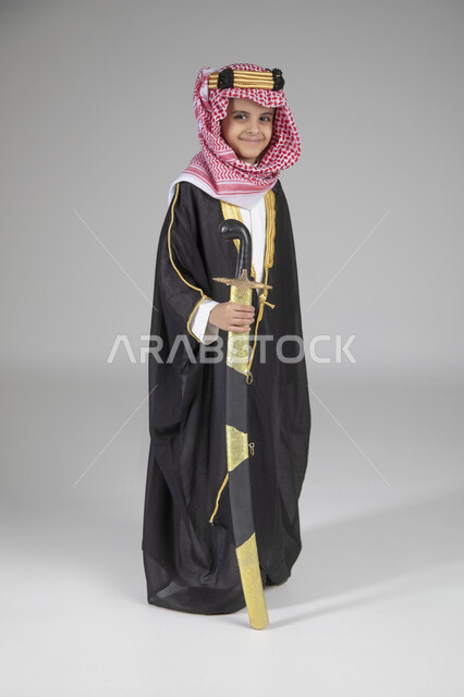  إحياء ذكرى يوم التأسيس والاحتفال بالمناسبات الوطنية الشعبية، بورتريه لولد عربي خليجي سعودي يرتدي البشت الأسود والعقال الذهبي العريض يمسك السيف بيده وينظر إلى الكاميرا بإيماءات العز والفخر، ارتداء الأزياء التقليدية التراثية، الجسم بالطول الكامل، خلفية رمادية
