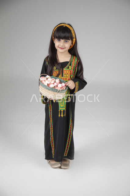 توزيع الحلويات والضيافة في المناسبات الوطنية، بورتريه لبنت عربية خليجية سعودية ترتدي الزي المطرز التقليدي السعودي النسائي تحمل سلة من الحلوى وتنظر إلى الكاميرا بإيماءات السعادة، ارتداء الأزياء التراثية لإحياء ذكرى يوم التأسيس، الجسم بالطول الكامل، خلفية رمادية