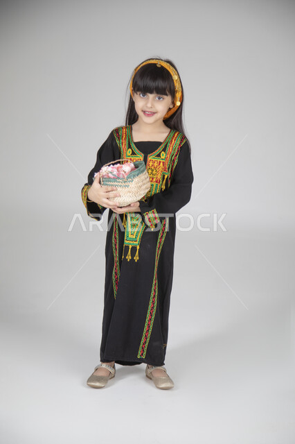 توزيع الحلويات والضيافة في المناسبات الوطنية، بورتريه لبنت عربية خليجية سعودية ترتدي الزي المطرز التقليدي السعودي النسائي تحمل سلة من الحلوى وتنظر إلى الكاميرا بإيماءات السعادة، ارتداء الأزياء التراثية لإحياء ذكرى يوم التأسيس، الجسم بالطول الكامل، خلفية رمادية