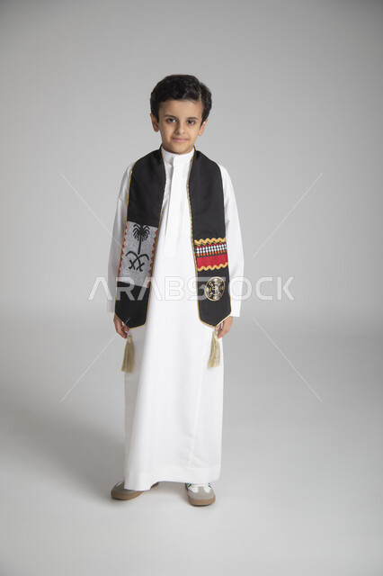 الاحتفال بذكرى يوم التأسيس، الأعياد الوطنية السنوية الشعبية، بورتريه لولد عربي خليجي سعودي يرتدي الثوب التقليدي ووشاح المملكة العربية السعودية ذو اللون الأسود بنقوش السدو، النظر إلى الكاميرا بإيماءات السعادة والسرور، الجسم بالطول الكامل، خلفية رمادية
