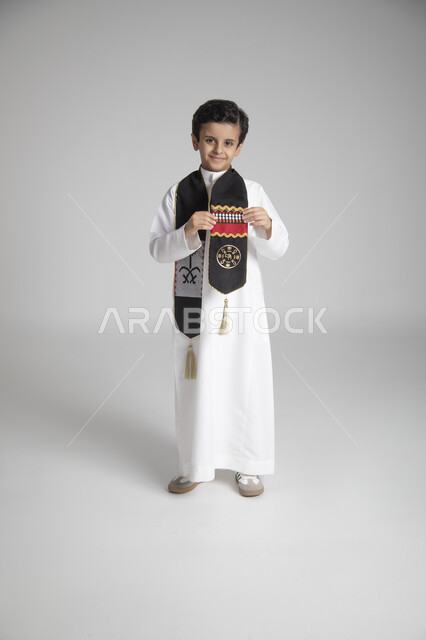 الاحتفال بذكرى يوم التأسيس، الأعياد الوطنية السنوية الشعبية، بورتريه لولد عربي خليجي سعودي يرتدي الثوب التقليدي ويمسك بوشاح المملكة العربية السعودية ذو اللون الأسود بنقوش السدو، النظر إلى الكاميرا بإيماءات السعادة والسرور، الجسم بالطول الكامل، خلفية رمادية