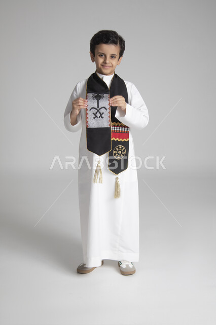 الاحتفال بذكرى يوم التأسيس، الأعياد الوطنية السنوية الشعبية، بورتريه لولد عربي خليجي سعودي يرتدي الثوب التقليدي ويمسك بوشاح المملكة العربية السعودية ذو اللون الأسود بنقوش السدو، النظر إلى الكاميرا بإيماءات السعادة والسرور، الجسم بالطول الكامل، خلفية رمادية