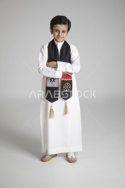 الاحتفال بذكرى يوم التأسيس، الأعياد الوطنية السنوية الشعبية، بورتريه لولد عربي خليجي سعودي يرتدي الثوب التقليدي ووشاح المملكة العربية السعودية ذو اللون الأسود بنقوش السدو، النظر إلى الكاميرا بإيماءات السعادة والسرور، الجسم بالطول الكامل، خلفية رمادية