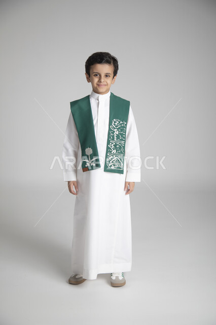 Standing with pride, annual national holidays, celebrating the founding day, portrait of a Saudi Arabian Gulf boy wearing the traditional dress and green Saudi Arabia scarf, looking at the camera with happy gestures, full body, gray background