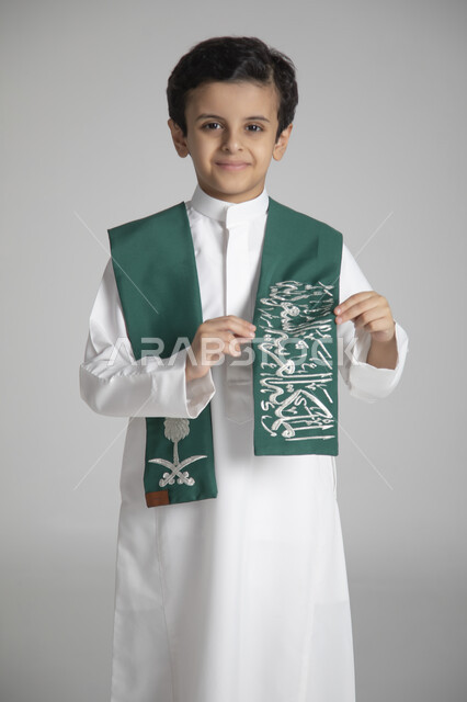 الأعياد الوطنية السنوية الشعبية، الاحتفال بذكرى يوم التأسيس، بورتريه مقرب لولد عربي خليجي سعودي يرتدي الثوب التقليدي ويمسك بوشاح المملكة العربية السعودية ذو اللون الأخضر بمشاعر الفخر والاعتزاز، النظر إلى الكاميرا بإيماءات السعادة والسرور، خلفية رمادية