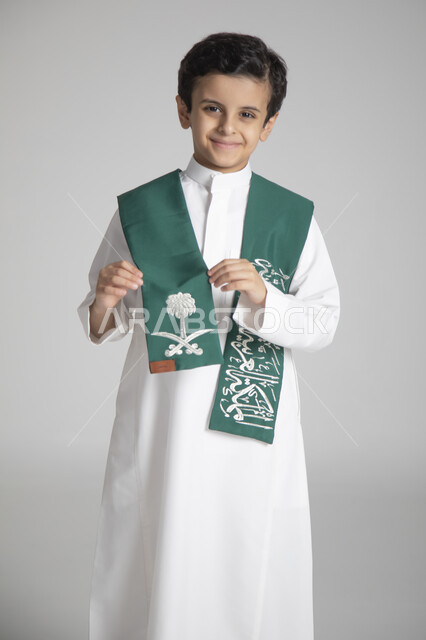 الأعياد الوطنية السنوية الشعبية، الاحتفال بذكرى يوم التأسيس، بورتريه مقرب لولد عربي خليجي سعودي يرتدي الثوب التقليدي ويمسك بوشاح المملكة العربية السعودية ذو اللون الأخضر بمشاعر الفخر والاعتزاز، النظر إلى الكاميرا بإيماءات السعادة والسرور، خلفية رمادية