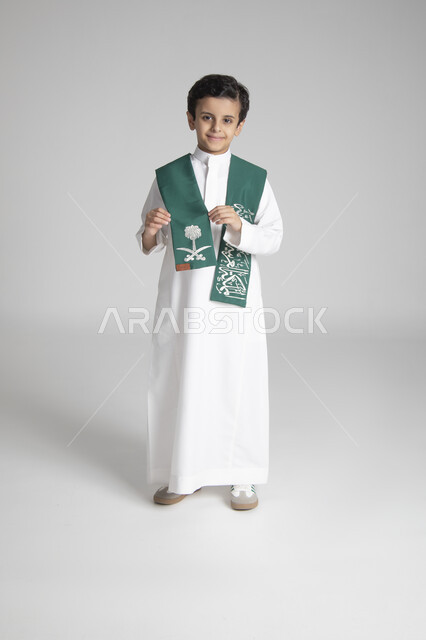 الأعياد الوطنية السنوية الشعبية، الاحتفال بذكرى يوم التأسيس، بورتريه لولد عربي خليجي سعودي يرتدي الثوب التقليدي ويمسك بوشاح المملكة العربية السعودية ذو اللون الأخضر بمشاعر الفخر، النظر إلى الكاميرا بإيماءات السعادة والسرور، الجسم بالطول الكامل، خلفية رمادية