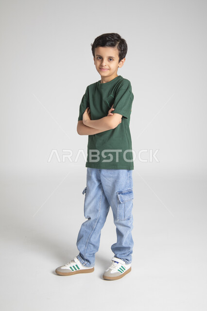 الوقوف بأيدي متقاطعة والنظر للكاميرا بإيماءات السرور، إحياء ذكرى يوم العلم السعودي، المناسبات والأعياد الشعبية الوطنية، تربية الأبناء على حب الوطن، بورتريه لولد عربي خليجي سعودي يرتدي تيشرت خاص للاحتفال باليوم الوطني السعودي، الجسم بالطول الكامل، خلفية رمادية