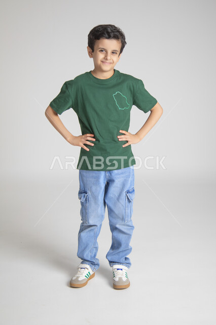 النظر للكاميرا بإيماءات السرور والسعادة، إحياء ذكرى يوم العلم السعودي، المناسبات والأعياد الشعبية الوطنية، تربية الأبناء على حب الوطن والانتماء له، بورتريه لولد عربي خليجي سعودي يرتدي تيشرت خاص للاحتفال باليوم الوطني السعودي، الجسم بالطول الكامل، خلفية رمادية