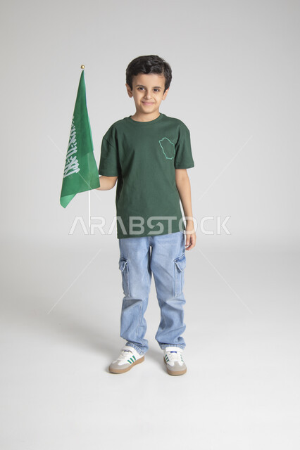 Raising the Kingdom's flag and looking at the camera with gestures of pleasure, commemorating Saudi Flag Day, national popular occasions and holidays, raising children to love the homeland, portrait of a Saudi Gulf Arab boy wearing a special T-shirt to celebrate Saudi National Day, full body, gray background