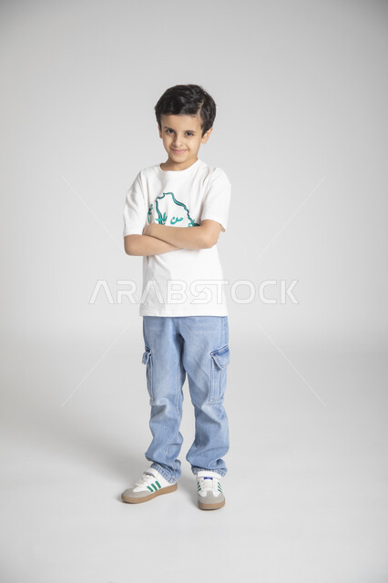 الوقوف بأيدي متقاطعة والنظر للكاميرا بإيماءات السرور، إحياء ذكرى يوم العلم السعودي، المناسبات والأعياد الشعبية الوطنية، تربية الأبناء على حب الوطن، بورتريه لولد عربي خليجي سعودي يرتدي تيشرت خاص للاحتفال باليوم الوطني السعودي، الجسم بالطول الكامل، خلفية رمادية