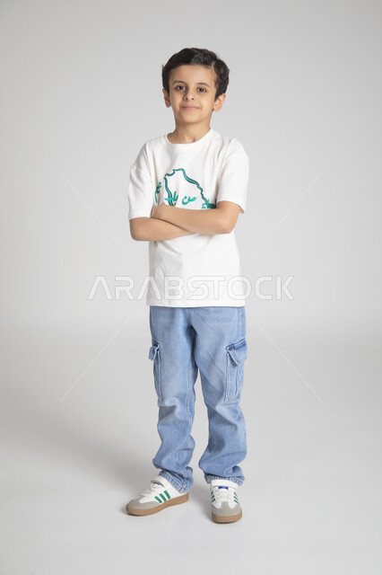 الوقوف بأيدي متقاطعة والنظر للكاميرا بإيماءات السرور، إحياء ذكرى يوم العلم السعودي، المناسبات والأعياد الشعبية الوطنية، تربية الأبناء على حب الوطن، بورتريه لولد عربي خليجي سعودي يرتدي تيشرت خاص للاحتفال باليوم الوطني السعودي، الجسم بالطول الكامل، خلفية رمادية