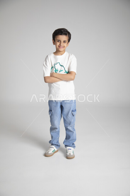 الوقوف بأيدي متقاطعة والنظر للكاميرا بإيماءات السرور، إحياء ذكرى يوم العلم السعودي، المناسبات والأعياد الشعبية الوطنية، تربية الأبناء على حب الوطن، بورتريه لولد عربي خليجي سعودي يرتدي تيشرت خاص للاحتفال باليوم الوطني السعودي، الجسم بالطول الكامل، خلفية رمادية