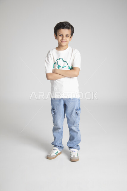 الوقوف بأيدي متقاطعة والنظر للكاميرا بإيماءات السرور، إحياء ذكرى يوم العلم السعودي، المناسبات والأعياد الشعبية الوطنية، تربية الأبناء على حب الوطن، بورتريه لولد عربي خليجي سعودي يرتدي تيشرت خاص للاحتفال باليوم الوطني السعودي، الجسم بالطول الكامل، خلفية رمادية