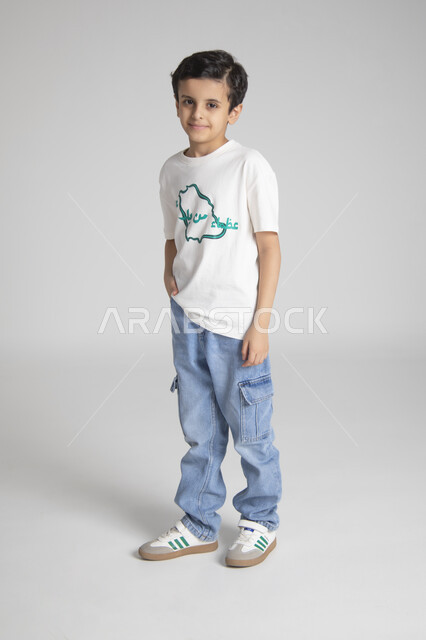 النظر للكاميرا بإيماءات السرور والسعادة، إحياء ذكرى يوم العلم السعودي، المناسبات والأعياد الشعبية الوطنية، تربية الأبناء على حب الوطن والانتماء له، بورتريه لولد عربي خليجي سعودي يرتدي تيشرت خاص للاحتفال باليوم الوطني السعودي، الجسم بالطول الكامل، خلفية رمادية