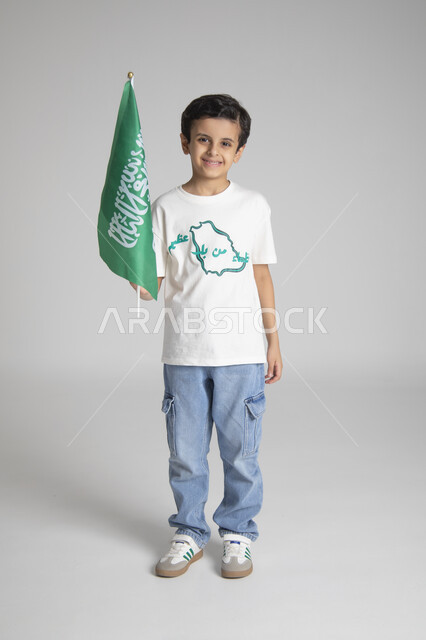 إحياء ذكرى يوم العلم السعودي، الاحتفالات بالمناسبات والأعياد الشعبية الوطنية، تربية الأبناء على حب الوطن، بورتريه لولد عربي خليجي سعودي يرتدي تيشرت خاص للاحتفال باليوم الوطني السعودي ويحمل راية المملكة العربية السعودية، الجسم بالطول الكامل، خلفية رمادية