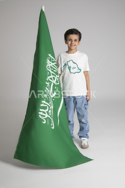 إحياء ذكرى يوم العلم السعودي، الاحتفالات بالمناسبات والأعياد الشعبية الوطنية، تربية الأبناء على حب الوطن، بورتريه لولد عربي خليجي سعودي يرتدي تيشرت خاص للاحتفال باليوم الوطني السعودي ويحمل راية المملكة العربية السعودية، الجسم بالطول الكامل، خلفية رمادية