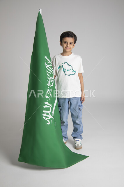 إحياء ذكرى يوم العلم السعودي، الاحتفالات بالمناسبات والأعياد الشعبية الوطنية، تربية الأبناء على حب الوطن، بورتريه لولد عربي خليجي سعودي يرتدي تيشرت خاص للاحتفال باليوم الوطني السعودي ويحمل راية المملكة العربية السعودية، الجسم بالطول الكامل، خلفية رمادية