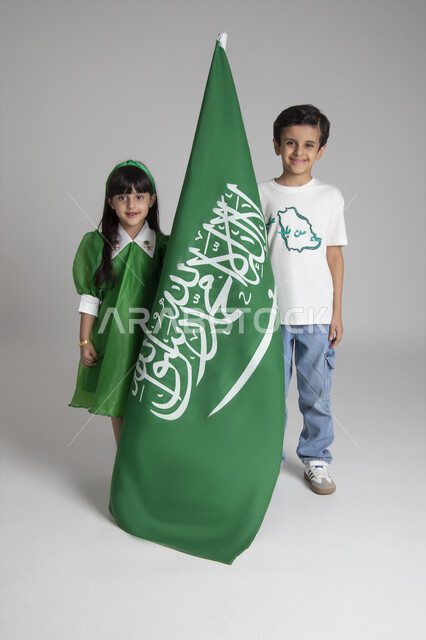 الاحتفال بيوم العلم السعودي، إحياء ذكرى المناسبات والأعياد الشعبية الوطنية، تربية الأبناء على حب الوطن، بورتريه لبنت وولد عربيين خليجيين سعوديين يرتديان ملابس خاصة باليوم الوطني السعودي ويرفعان راية السعودية بإيماءات الفخر، الجسم بالطول الكامل، خلفية رمادية