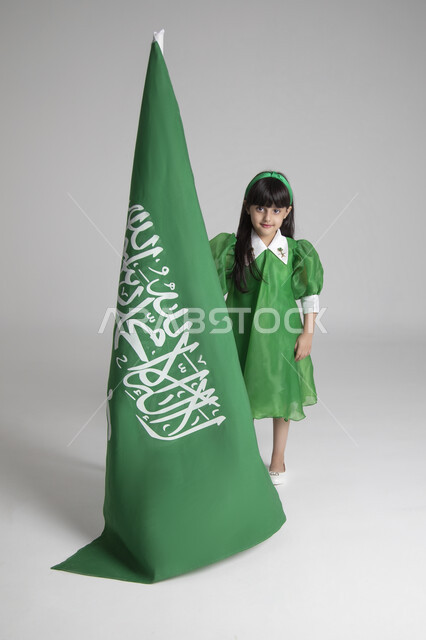 الاحتفال بيوم العلم السعودي، إحياء ذكرى المناسبات والأعياد الشعبية الوطنية، تربية الأبناء على حب الوطن، بورتريه لبنت عربية خليجية سعودية ترتدي ثوب أخضر اللون ابتهاجا باليوم الوطني السعودي ترفع راية السعودية بإيماءات الفخر، الجسم بالطول الكامل، خلفية رمادية