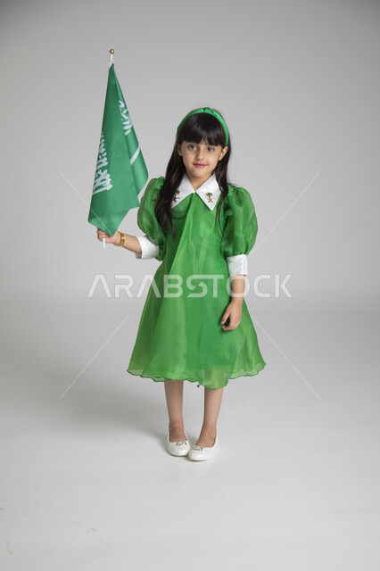 الاحتفال بيوم العلم السعودي، إحياء ذكرى المناسبات والأعياد الشعبية الوطنية، تربية الأبناء على حب الوطن، بورتريه لبنت عربية خليجية سعودية ترتدي ثوب أخضر اللون ابتهاجا باليوم الوطني السعودي ترفع راية السعودية بإيماءات الفخر، الجسم بالطول الكامل، خلفية رمادية