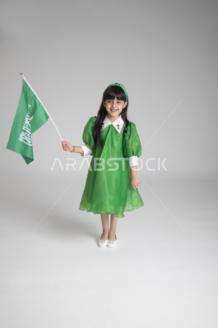 إحياء ذكرى المناسبات والأعياد الشعبية الوطنية، تربية الأبناء على حب الوطن، بورتريه لبنت عربية خليجية سعودية ترتدي ثوب أخضر اللون ابتهاجا باليوم الوطني السعودي ترفع راية السعودية بإيماءات الفخر، الجسم بالطول الكامل، الاحتفال بيوم العلم السعودي، خلفية رمادية