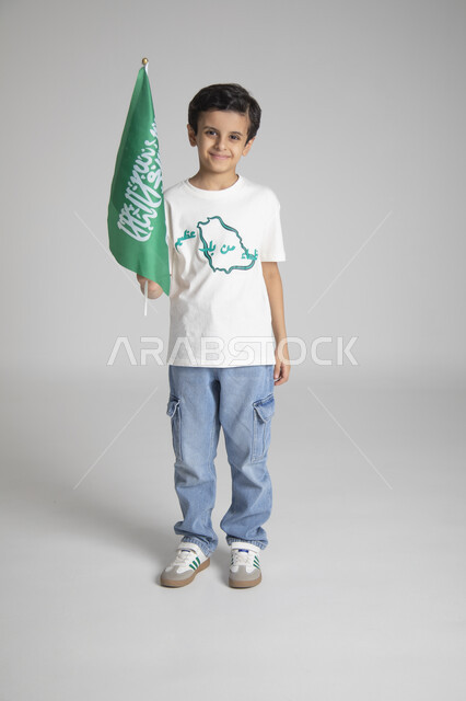 إحياء ذكرى يوم العلم السعودي، الاحتفالات بالمناسبات والأعياد الشعبية الوطنية، تربية الأبناء على حب الوطن، بورتريه لولد عربي خليجي سعودي يرتدي تيشرت خاص للاحتفال باليوم الوطني السعودي ويحمل راية المملكة العربية السعودية، الجسم بالطول الكامل، خلفية رمادية
