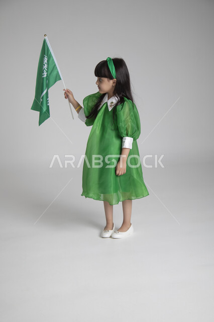 إحياء ذكرى المناسبات والأعياد الشعبية الوطنية، تربية الأبناء على حب الوطن، بورتريه لبنت عربية خليجية سعودية ترتدي ثوب أخضر اللون ابتهاجا باليوم الوطني السعودي ترفع راية السعودية بإيماءات الفخر، الجسم بالطول الكامل، الاحتفال بيوم العلم السعودي، خلفية رمادية