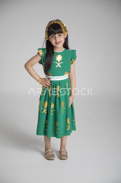 إحياء ذكرى المناسبات والأعياد الشعبية الوطنية، تربية الأبناء على حب الوطن، بورتريه لبنت عربية خليجية سعودية ترتدي ثوب أخضر اللون ابتهاجا باليوم الوطني السعودي تنظر للكاميرا بإيماءات السرور، الجسم بالطول الكامل، الاحتفال بيوم العلم السعودي بفخر، خلفية رمادية