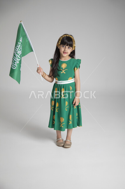 إحياء ذكرى المناسبات والأعياد الشعبية الوطنية، تربية الأبناء على حب الوطن، بورتريه لبنت عربية خليجية سعودية ترتدي ثوب أخضر اللون ابتهاجا باليوم الوطني السعودي ترفع راية السعودية بإيماءات الفخر، الجسم بالطول الكامل، الاحتفال بيوم العلم السعودي، خلفية رمادية
