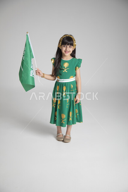 إحياء ذكرى المناسبات والأعياد الشعبية الوطنية، تربية الأبناء على حب الوطن، بورتريه لبنت عربية خليجية سعودية ترتدي ثوب أخضر اللون ابتهاجا باليوم الوطني السعودي ترفع راية السعودية بإيماءات الفخر، الجسم بالطول الكامل، الاحتفال بيوم العلم السعودي، خلفية رمادية
