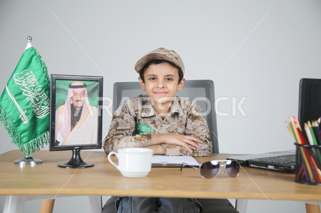 تجسيد المهن المستقبلية للأطفال، بورتريه لولد عربي خليجي سعودي يرتدي زي الشرطة يجلس خلف المكتب بجانبه العلم السعودي وصورة لملك المملكة العربية السعودية، النظر إلى الكاميرا بإيماءات السرور، اختيار مهنة الضابط الشرطي والعمل في المجال العسكري،  خلفية رمادية