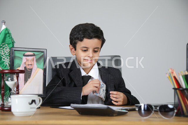 العمل في المجال الإداري والمكتبي واختيار مهنة المحاسب، تجسيد المهن المستقبلية للأطفال، بورتريه مقرب لولد عربي خليجي سعودي يرتدي البدلة الرسمية يجلس خلف المكتب وبجانبه العلم السعودي وصورة لملك السعودية، الأوراق والمستندات والحاسبة الآلية الرقمية، خلفية رمادية