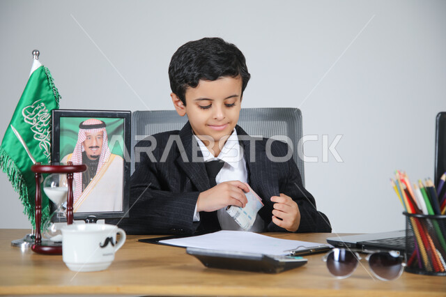 العمل في المجال الإداري والمكتبي واختيار مهنة المحاسب، تجسيد المهن المستقبلية للأطفال، بورتريه مقرب لولد عربي خليجي سعودي يرتدي البدلة الرسمية يجلس خلف المكتب وبجانبه العلم السعودي وصورة لملك السعودية، الأوراق والمستندات والحاسبة الآلية الرقمية، خلفية رمادية