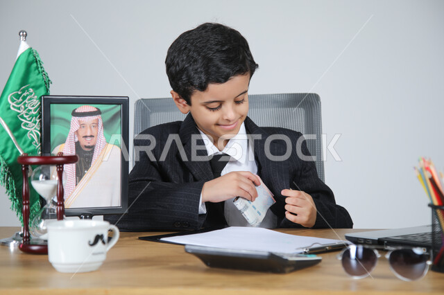 العمل في المجال الإداري والمكتبي واختيار مهنة المحاسب، تجسيد المهن المستقبلية للأطفال، بورتريه مقرب لولد عربي خليجي سعودي يرتدي البدلة الرسمية يجلس خلف المكتب وبجانبه العلم السعودي وصورة لملك السعودية، الأوراق والمستندات والحاسبة الآلية الرقمية، خلفية رمادية