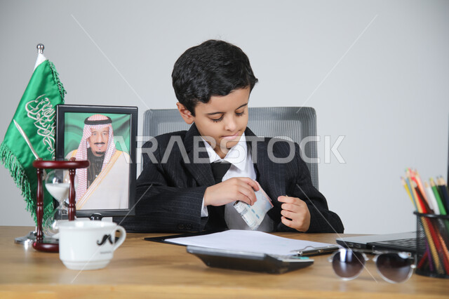 اختيار مهنة المحاسب والعمل في المجال الإداري والمكتبي، تجسيد المهن المستقبلية للأطفال، بورتريه لولد عربي خليجي سعودي يرتدي البدلة الرسمية يجلس خلف المكتب وبجانبه العلم السعودي وصورة لملك السعودية، الأوراق والمستندات والحاسبة الآلية على الطاولة، خلفية رمادية