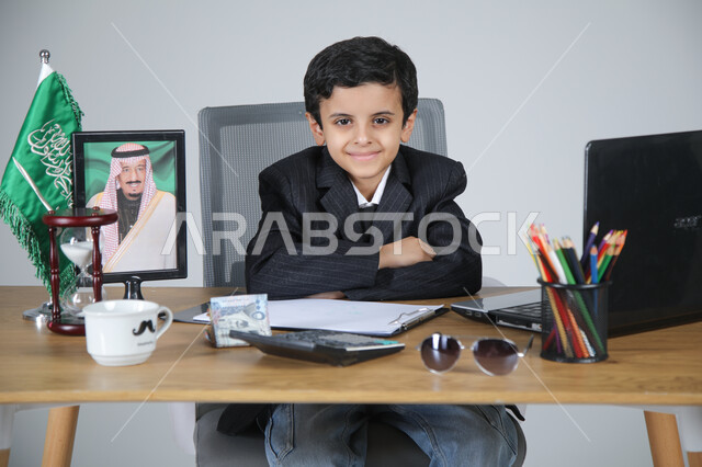 اختيار مهنة المحاسب والعمل في المجال الإداري والمكتبي، تجسيد المهن المستقبلية للأطفال، بورتريه لولد عربي خليجي سعودي يرتدي البدلة الرسمية يجلس خلف المكتب وبجانبه العلم السعودي وصورة لملك السعودية، الأوراق والمستندات والحاسبة الآلية على الطاولة، خلفية رمادية