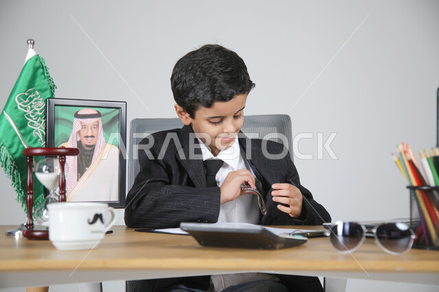 اختيار مهنة المحاسب والعمل في المجال الإداري والمكتبي، تجسيد المهن المستقبلية للأطفال، بورتريه لولد عربي خليجي سعودي يرتدي البدلة الرسمية يجلس خلف المكتب وبجانيه العلم السعودي وصورة لملك المملكة العربية السعودية، المستندات والورق والحاسبة الرقمية، خلفية رمادية