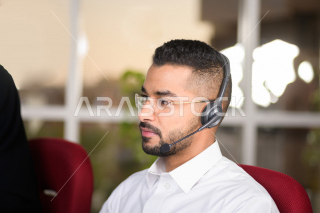 موظف عربي خليجي سعودي يعمل في خدمة العملاء ويرتدي سماعة الاتصال، استعلامات واستفسارات، الاجابه على اسئلة العملاء، اجراء اتصال فيديو من خلال الحاسوب، نظارة طبية، متابعة وتطوير العمل