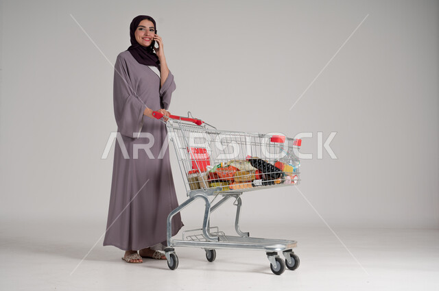 إجراء مكالمة هاتفية عبر الجوال، الاستمتاع بالتسوق في المولات والمراكز التجارية، الاستفادة من الخصومات على المنتجات، بورتريه لامرأة عربية خليجية سعودية محجبة ترتدي العباءة تقف وتستخدم الهاتف المحمول وأمامها عربة المشتريات، الجسم بالطول الكامل، خلفية رمادية