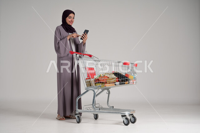 استخدام الهاتف المحمول أثناء التسوق في المولات والمراكز التجارية، الاستفادة من الخصومات والتنزيلات على المنتجات، بورتريه لامرأة عربية خليجية سعودية محجبة ترتدي العباءة التقليدية تقف وتمسك الجوال بيدها وأمامها عربة المشتريات، الجسم بالطول الكامل، خلفية رمادية